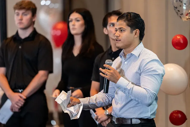 Man in light blue shirt holds microphone while speaking, flanked by colleagues in black and dark clothing.