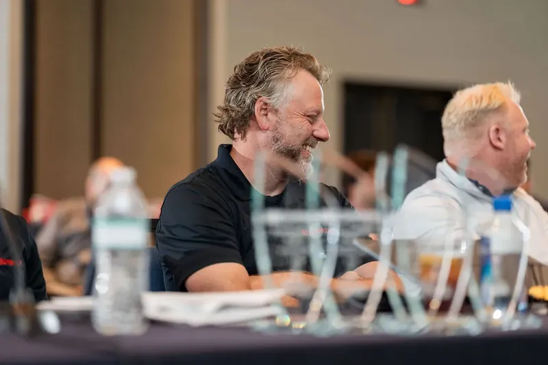 Man with dark beard in black shirt speaks at panel table with water bottles and colleagues seated nearby.