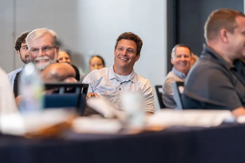 Man in light shirt smiles widely at table during event, surrounded by other attendees in casual business attire.
