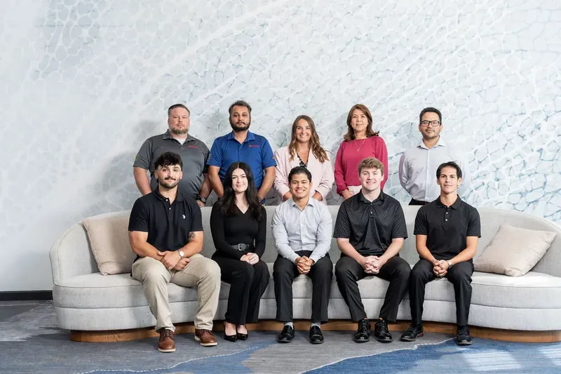 Nine team members sit and stand together against a white textured wall, wearing casual attire in neutrals and jewel tones.