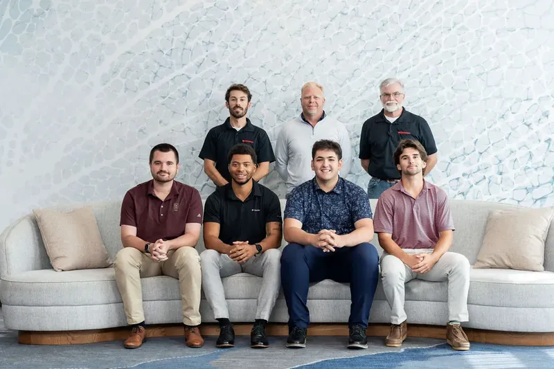 Eight men pose on cream-colored seating against a mottled gray and white backdrop, dressed in casual shirts in varied colors.