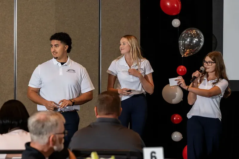 Two presenters in white company polos stand before an audience while a colleague holds colorful decorative spheres on a black