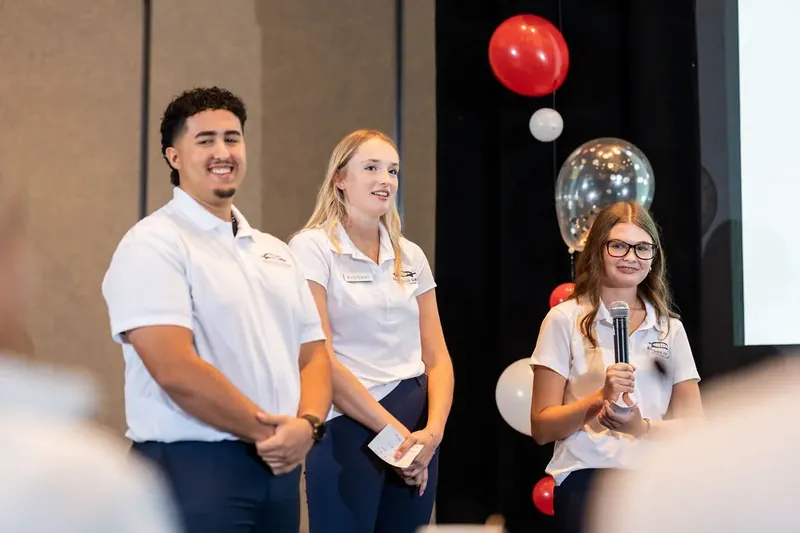 Three team members in white polos speak to seated attendees, with hanging red and metallic balloons visible behind them.
