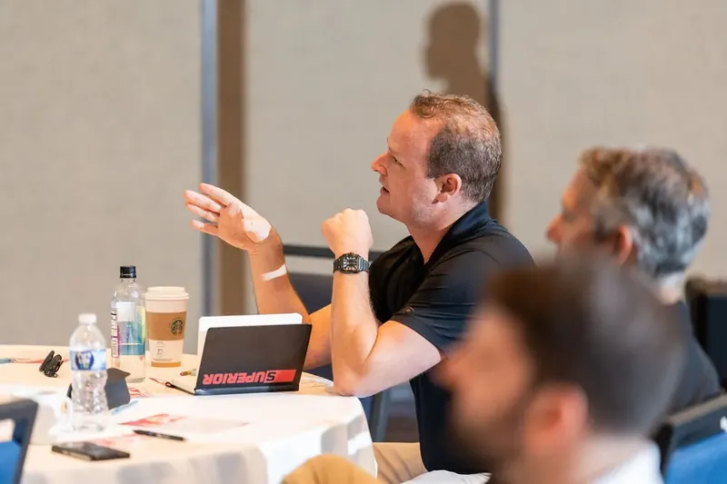 Man in black polo shirt gestures while speaking at a conference table with colleagues and water bottles nearby.