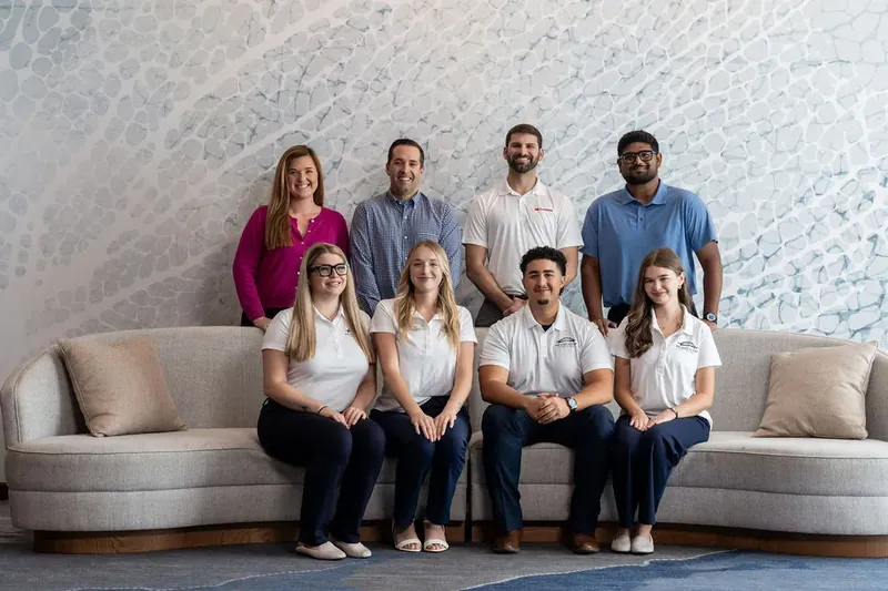 Nine team members pose together on a curved gray sofa against a brick wall.