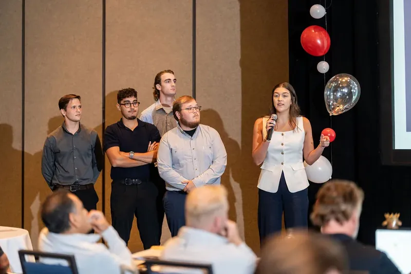Woman in white vest and orange bowling ball stands with four colleagues during an indoor presentation.