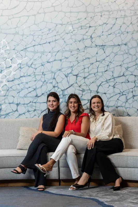 Three women sit on a gray sofa against a textured blue-and-white wall backdrop.