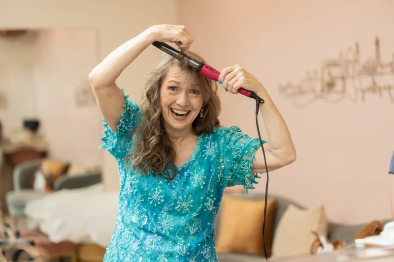 Sybil smiles while curling her hair with a styling iron in a brightly lit preparation room.