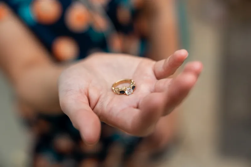A gold engagement ring with a square stone rests in an open palm, with blurred figures in patterned clothing behind.