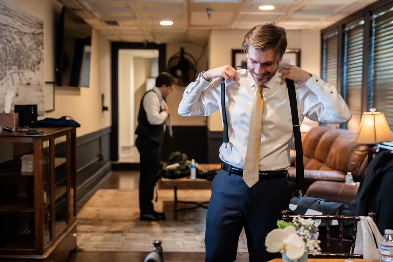 Chris adjusts his blue tie and vest while getting ready in an elegantly furnished interior space.