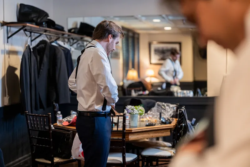 Chris adjusts his tie with both hands while smiling in a warm-lit indoor space.