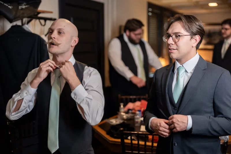 Two men in suits adjust their neckties side by side in an elegant venue hallway.