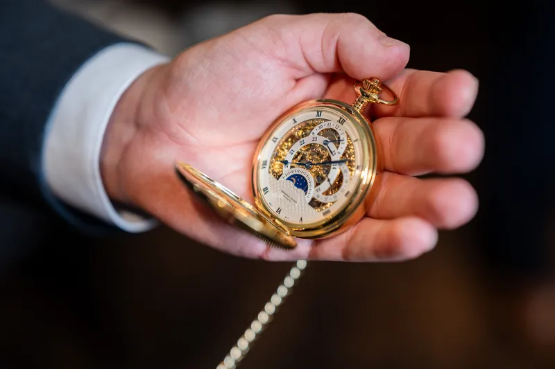 A hand in a white dress shirt cuff holds a gold pocket watch with a pearl chain.