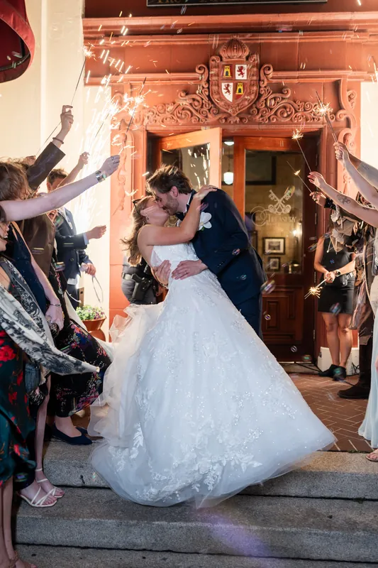 Sybil exits the venue in her layered white dress while guests wave enthusiastically from the entrance.