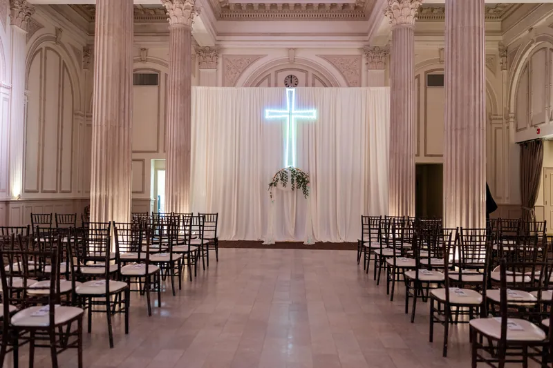 Black chiavari chairs line both sides of a symmetrical chapel with glowing cross and soaring columns.