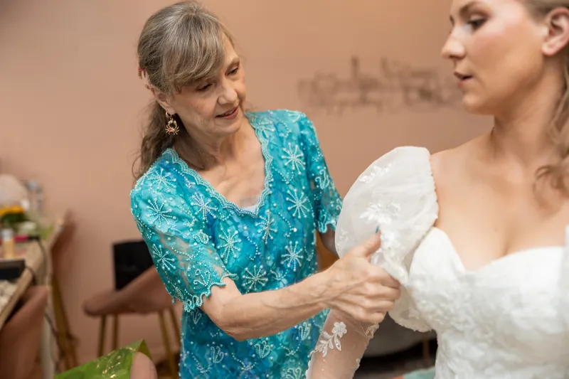 A woman in a turquoise patterned shirt adjusts Sybil's white lace wedding dress inside The Treasury on the Plaza.