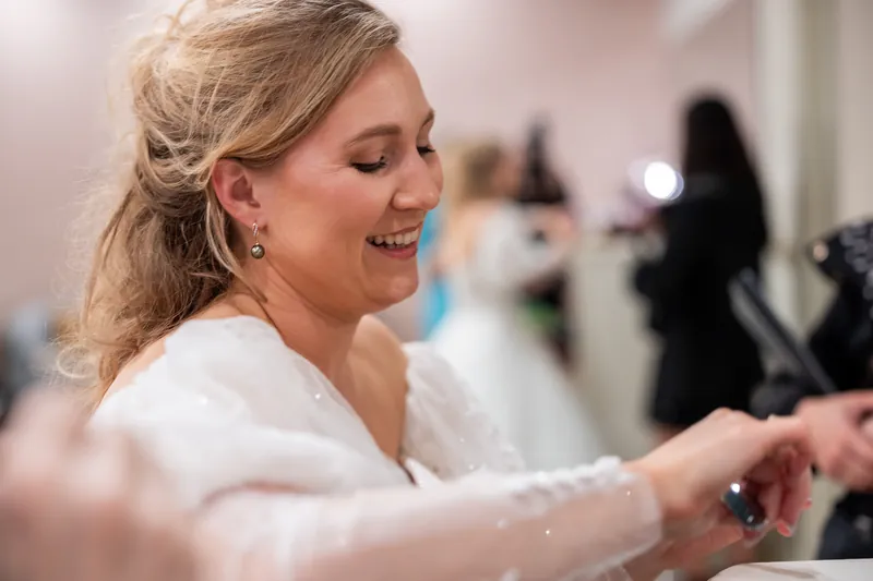 Sybil smiles while having her dress fastened, wearing a white off-shoulder gown in an indoor bridal preparation space.