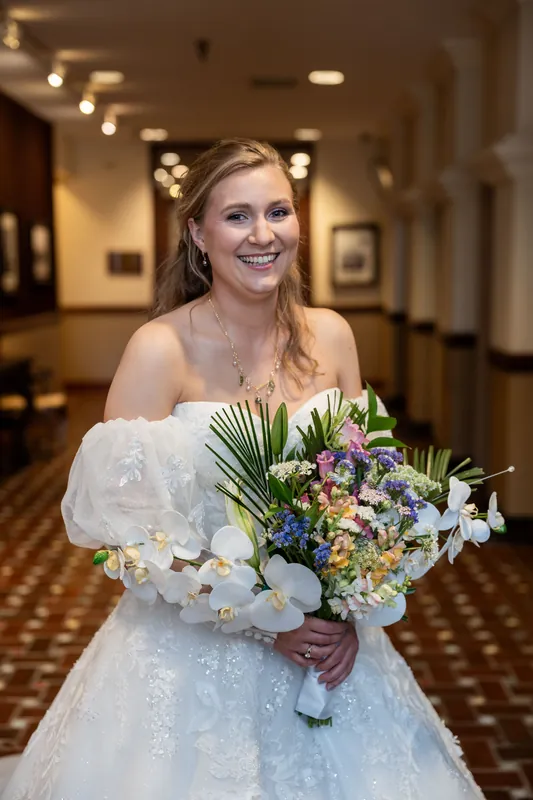 Sybil smiles while holding a lush bouquet of white orchids, purple flowers, and greenery inside The Treasury on the Plaza.