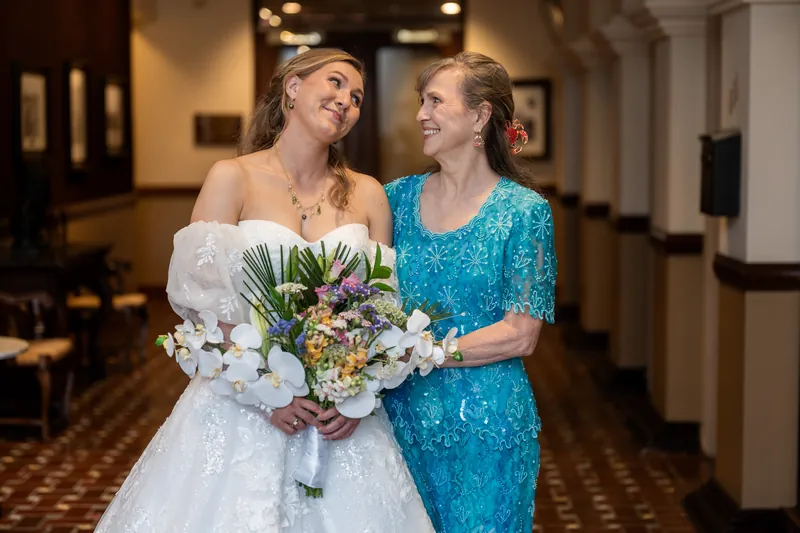 Sybil and a companion in a turquoise sequin dress walk together down an elegant hallway, both holding the wedding bouquet.