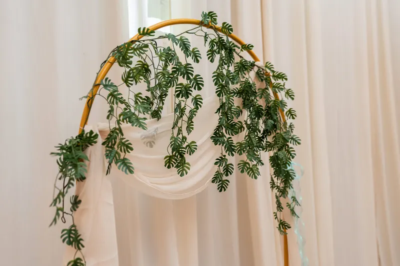 A brass arch draped with cascading ferns and greenery stands against sheer cream fabric at the venue.