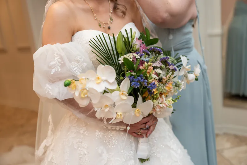 Sybil's companion holds the bridal bouquet of white orchids and mixed florals beside her in a light blue dress.