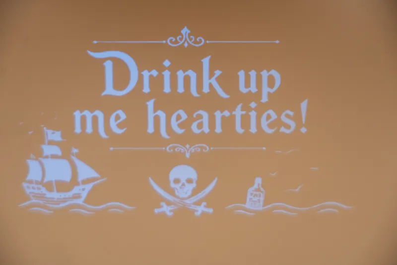 A wall projection reads "Drink up me hearties!" with pirate-themed graphics including a ship, skull, and bottle.