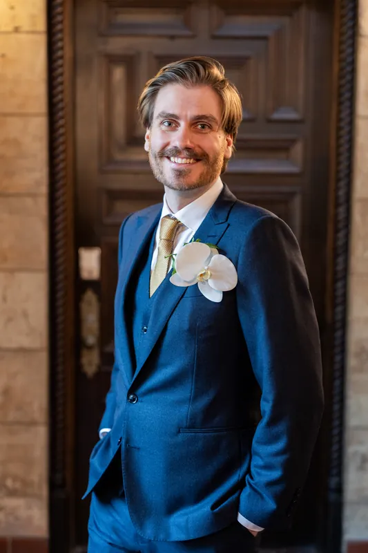 Chris stands smiling in a blue suit with a white boutonnière against an ornate wooden doorway at The Treasury on the Plaza.