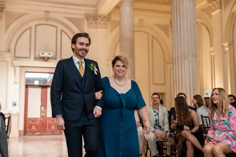 Chris and a family member walk arm-in-arm down a columned hall lined with seated guests.