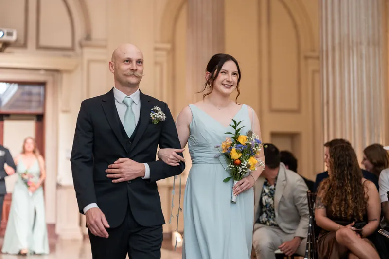 Sybil in a pale blue bridesmaid dress carries a yellow bouquet while walking with Chris through the historic venue.