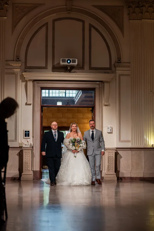 Sybil in a white wedding dress walks between Chris and another attendant under an arched doorway.