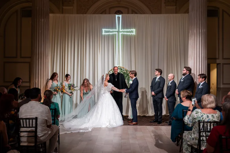 Sybil and Chris exchange vows beneath an illuminated cross as wedding guests look on inside the columned hall.