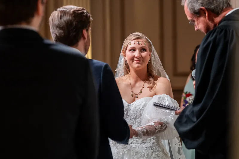 Sybil in an ornate lace gown smiles while surrounded by men in dark suits during a ceremony at The Treasury on the Plaza.