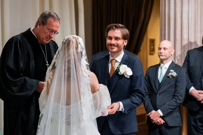 An officiant in black robes stands beside Chris in a navy suit as Sybil, veiled and in white lace, looks on during