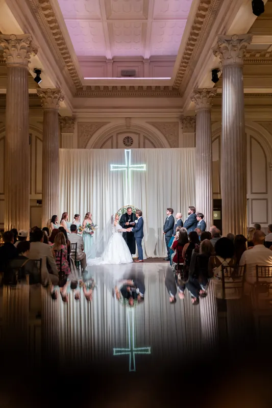 Sybil and Chris exchange vows beneath a soaring coffered ceiling and illuminated cross at The Treasury on the Plaza, guests