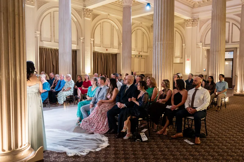 Wedding guests fill a grand columned hall with cream and beige furnishings as Sybil's train trails across polished flooring