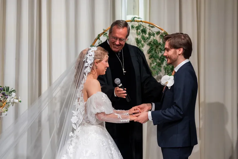 The officiant holds Sybil's hand as she and Chris face each other, veil flowing, in front of a floral arch draped with white