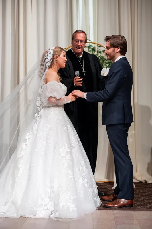 Sybil in her lace gown exchanges rings with Chris while the officiant reads vows, both standing on the ceremony floor between