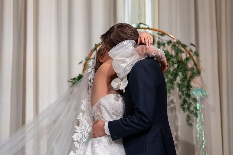 Sybil and Chris embrace under a floral arch with ivory curtains draped behind them at The Treasury on the Plaza.