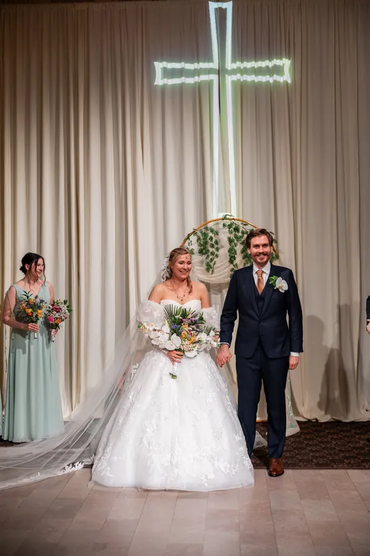 Sybil and Chris walk down the aisle together beneath a glowing cross, with a wedding party member visible to the left.