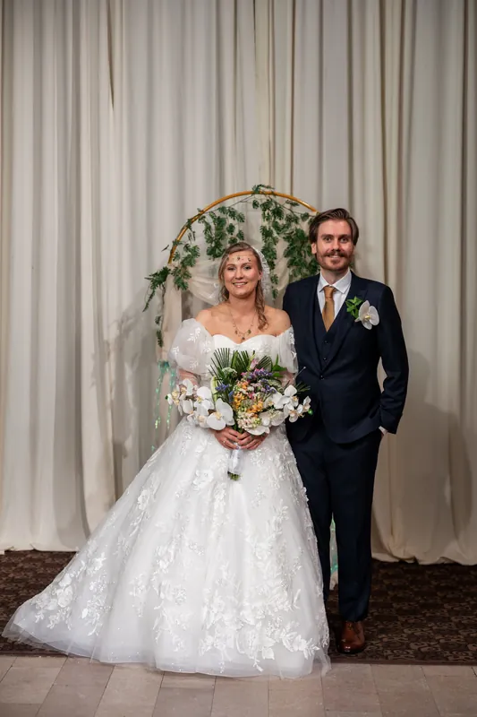 Sybil holds a white and gold bouquet while standing beside Chris in front of the draped floral backdrop.