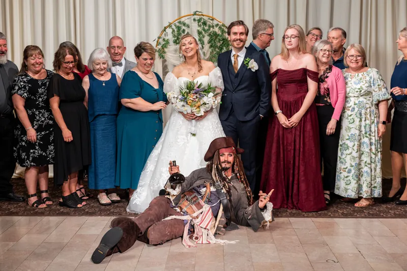 A large wedding party gathers for a group photo, with someone sitting cross-legged in front holding a small child.