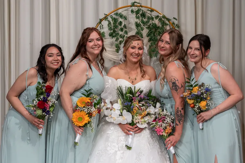 Sybil stands centered in a white gown surrounded by five attendants in sage green dresses, all holding colorful bouquets