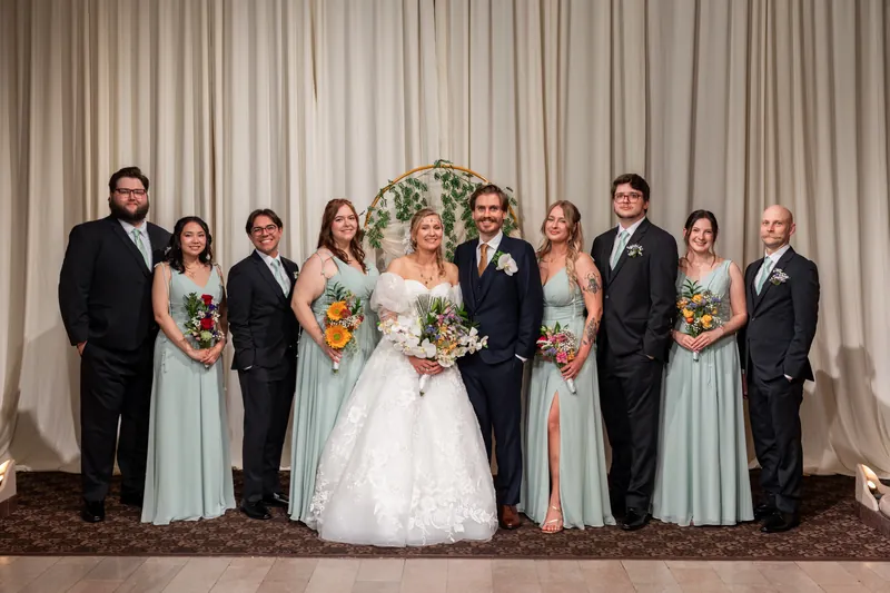 Sybil and Chris pose with their wedding party—five attendants in sage green and five groomsmen in black suits—beneath