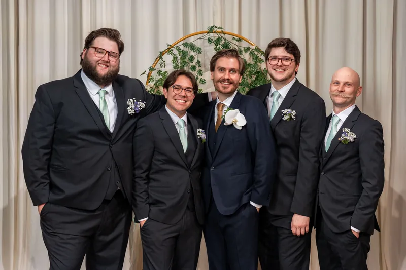 Chris stands with five groomsmen in dark suits and boutonnières, lined up against white draping at The Treasury on the Plaza.