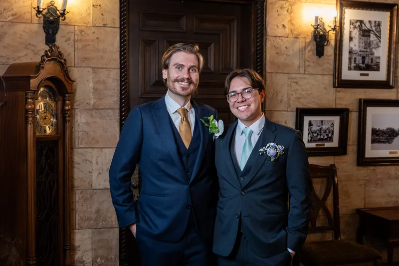 Chris and a groomsman in navy suits with white boutonnières stand in an ornate stone corridor with period sconces and framed