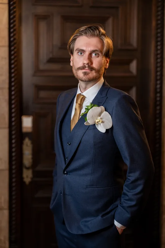 Chris in a navy blazer and gold tie adjusts his white orchid boutonnière while posing in a doorway at The Treasury