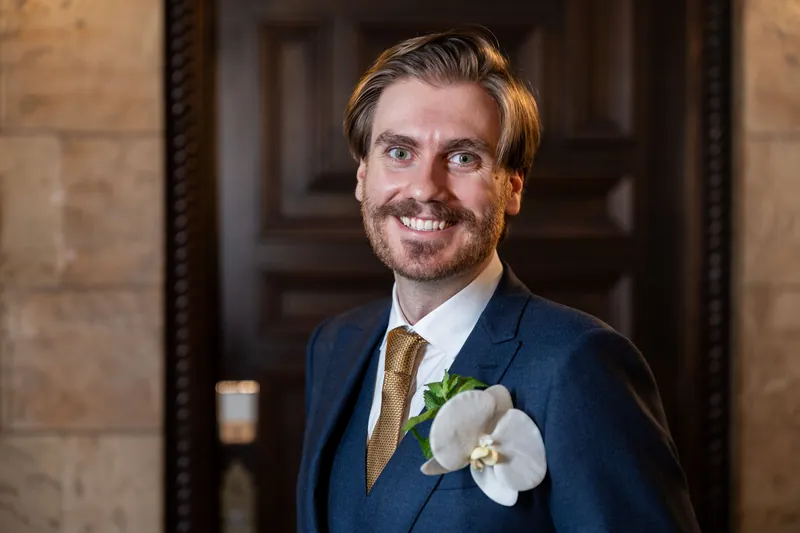 Chris smiles in a navy blazer and gold tie, his white orchid boutonnière visible as he stands in a stone-walled corridor.