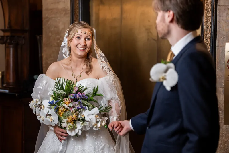 Sybil holds her bouquet while smiling at Chris in The Treasury's elegant interior, both dressed formally for their wedding.