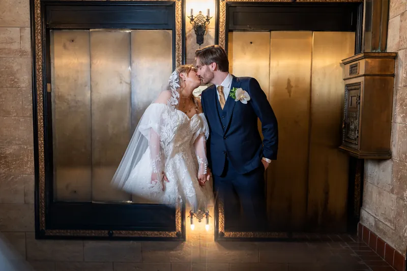 Sybil and Chris kiss by the elevator doors at The Treasury, with ornate gold panels framing the moment.