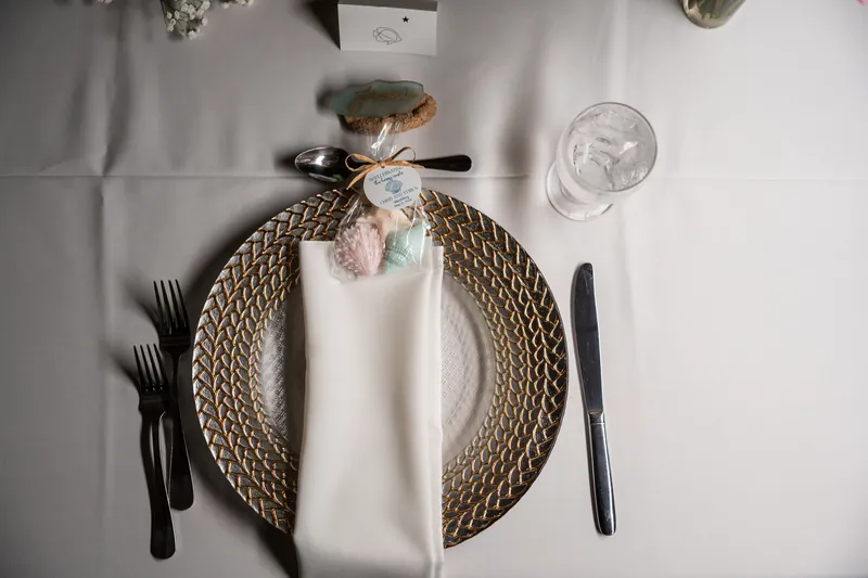 A place setting features a woven charger, white napkin, and small floral accent tied with ribbon on a neutral tablecloth.